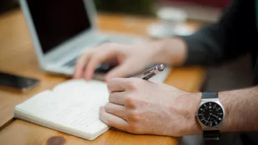 Pessoa escrevendo em um caderno com um laptop ao lado.