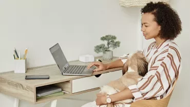 Mulher trabalha no notebook com cachorro no colo em um escritório em casa.