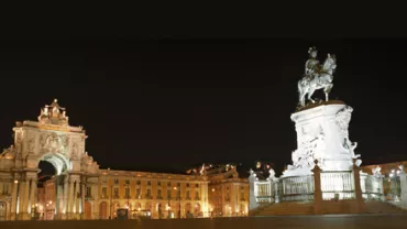 Praça iluminada à noite com estátua e arco ao fundo.