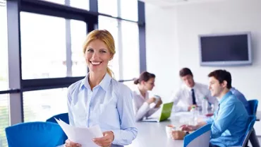 Mulher sorrindo com papéis na mão em sala de reunião.