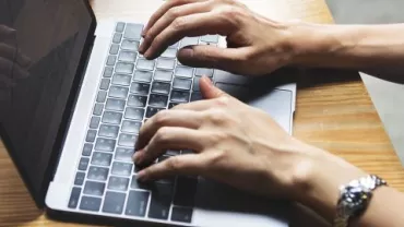 Mãos digitando em um laptop sobre uma mesa de madeira.